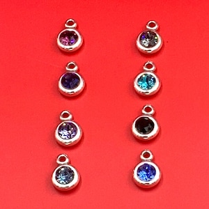 May include: Eight silver-toned pendants with colorful gemstones. The gemstones are in various colors, including purple, blue, and black. Each pendant has a small loop at the top for attaching to a chain or bracelet. The pendants are displayed against a red background.