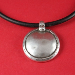 May include: A silver pendant with a round, smooth surface and a beaded border hangs from a black leather cord.
