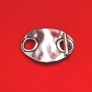 May include: A silver-toned toggle clasp featuring an oval-shaped plate with a hammered, textured surface. The clasp includes a circular loop on one side and a perpendicular bar attachment on the other, designed for securing jewelry or garment closures.