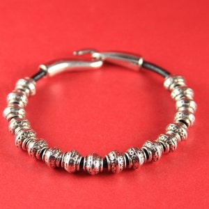 K&ouml;nnte beinhalten: Ein silbernes Armband mit Perlen und einem schwarzen Lederband mit einem silbernen Verschluss.