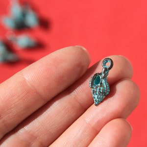 May include: A small, turquoise seashell charm with a dark metal loop for hanging. The charm is held in a hand against a red background, with other blurred charms visible.