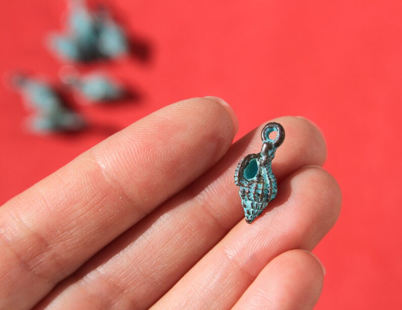 May include: A small, turquoise seashell charm with a dark metal loop for hanging. The charm is held in a hand against a red background, with other blurred charms visible.