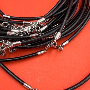 May include: A collection of black leather cord necklaces featuring polished silver-tone metal end caps and lobster claw clasps. The cords are arranged in a tangled pile, showcasing the smooth texture of the leather and the metallic finish of the hardware.
