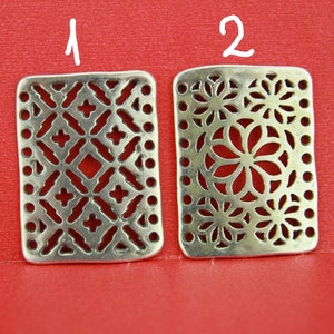 May include: Two silver metal charms with intricate cut-out designs. The charm on the left has a geometric pattern, while the charm on the right has a floral pattern.