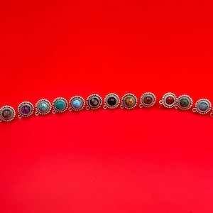 May include: A collection of silver-toned stud earrings with various colored gemstones, including blue, purple, green, and brown, arranged on a vibrant red background. Each earring features a circular gemstone surrounded by a decorative border.