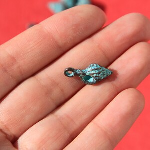May include: A small, turquoise-colored seashell pendant with a dark brown loop for hanging. The shell has detailed ridges and a textured surface. The pendant is held in a hand against a red background.