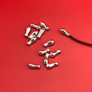 May include: A collection of small, shiny silver-colored beads scattered on a bright red surface. The beads have a teardrop shape with a rounded top and a narrower base. A black cord with a silver bead is visible.
