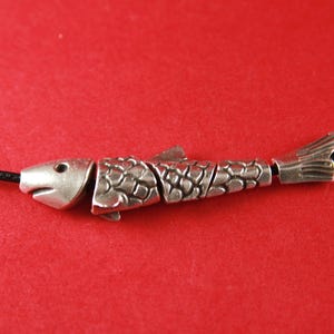 May include: A silver-toned fish-shaped pendant strung on a thin black cord. The pendant is segmented into four distinct pieces, featuring detailed scale patterns on the body, a defined head with an eye, and a flared tail fin at the end.
