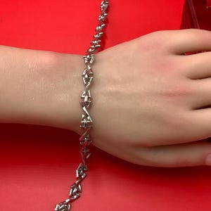 May include: A silver-toned metal chain bracelet featuring a series of repeating, angular, triangular-shaped links connected by small straight bars. The bracelet has a polished, reflective finish and is displayed against a solid, vibrant red background.