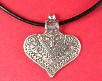 May include: A silver heart-shaped pendant with intricate designs hangs from a black leather cord. The pendant has a detailed, ornate pattern.