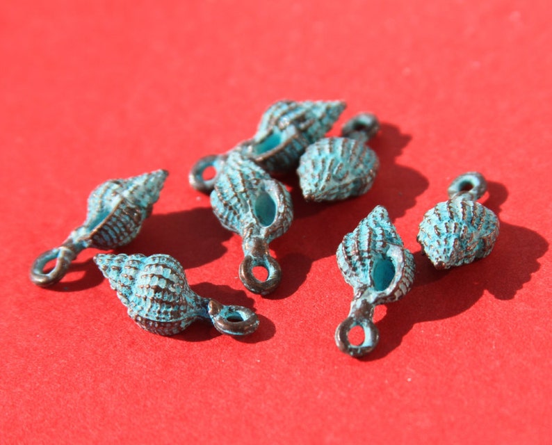 May include: Several small, turquoise-colored seashell charms with a bronze-colored finish are scattered on a red background. Each charm has a loop for attaching to jewelry or other crafts. The charms have a textured surface.