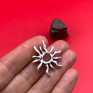 May include: A silver-colored sun pendant with a circular center and radiating rays. The pendant is held in a hand against a red background. A dark, rounded stone is visible in the background.