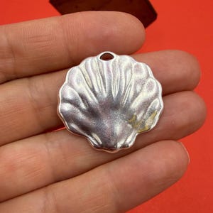 May include: A silver-colored, shell-shaped pendant with a small hole at the top. The pendant has a textured surface with radiating lines. A wooden, hexagonal shape is in the background against a red backdrop.