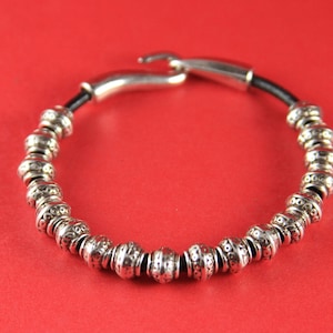 K&ouml;nnte beinhalten: Ein silbernes Armband mit Perlen und einem schwarzen Lederband und einem silbernen Verschluss. Die Perlen sind rund und haben ein strukturiertes Design.