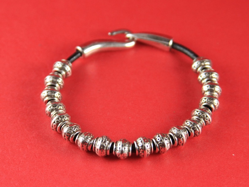 K&ouml;nnte beinhalten: Ein silbernes Armband mit Perlen und einem schwarzen Lederband und einem silbernen Verschluss. Die Perlen sind rund und haben ein strukturiertes Design.