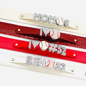 May include: Four white and red leather bracelets with rhinestones and baseball charms. The bracelets feature the words "MOM", "I LOVE", "#52", and "BEN#93".