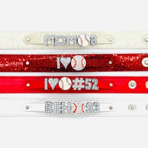 May include: Four sparkly bracelets with rhinestones and a baseball charm. The bracelets are white, red, and red glitter. The charms say "MOM", "I LOVE", "I LOVE #52", and "BEN#91".