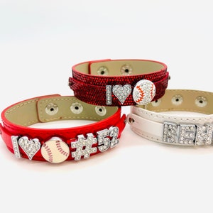 May include: Three red and white leather bracelets with rhinestones and charms. The charms include a heart, a baseball, a number 52, and the word "BENO".