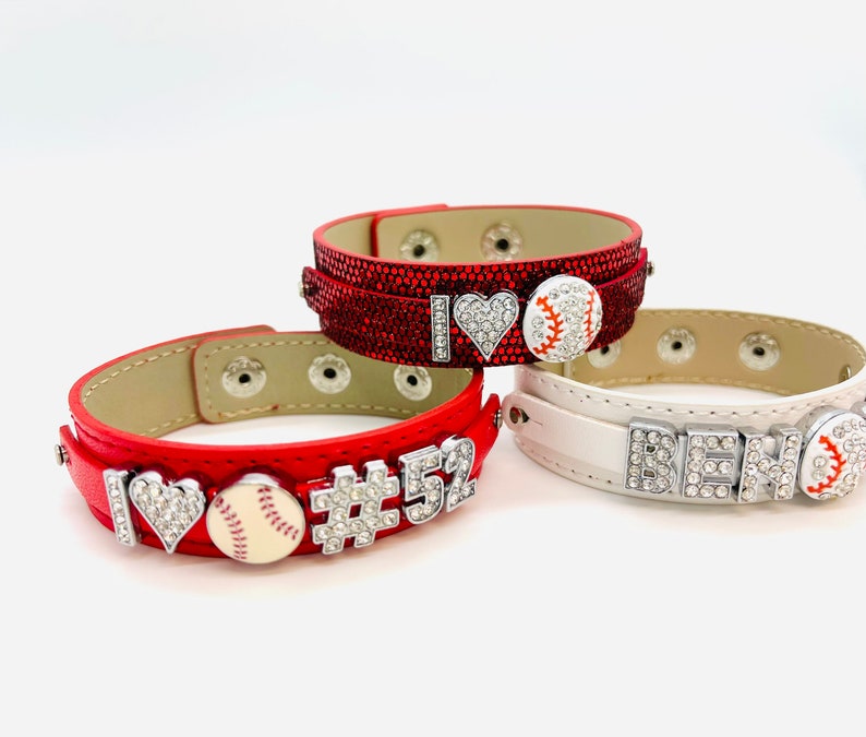 May include: Three red and white leather bracelets with rhinestones and charms. The charms include a heart, a baseball, a number 52, and the word "BENO".
