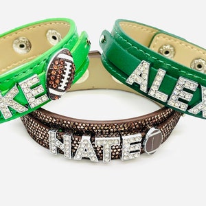 May include: Three green leather bracelets with rhinestones spelling out the names "LUKE", "ALEX", and "NATE". Each bracelet has a brown football charm.