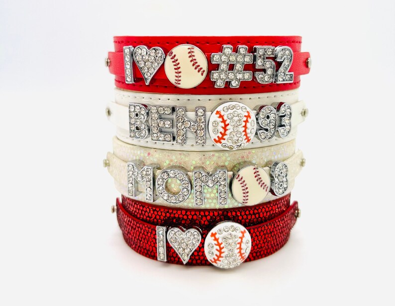 May include: Stack of four red and white leather bracelets with rhinestones and baseball charms. The bracelets feature the text "I love", "#52", "BEN", "MOM", and a baseball with rhinestones.