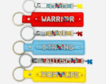 May include: Five colorful keychains with different words and a puzzle piece heart design. The keychains are red, yellow, blue, and silver. The words on the keychains are "MOM", "WARRIOR", "LOVE", "STRONG", and "AUTISM MOM LIFE".