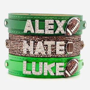 May include: Three green leather bracelets with rhinestones and football charms. The bracelets are stacked on top of each other. The top bracelet says "ALEX", the middle bracelet says "NATE", and the bottom bracelet says "LUKE".