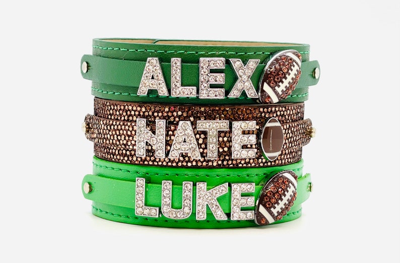 May include: Three green leather bracelets with rhinestones and football charms. The bracelets are stacked on top of each other. The top bracelet says "ALEX", the middle bracelet says "NATE", and the bottom bracelet says "LUKE".