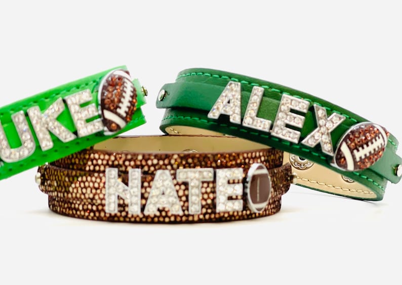 May include: Three leather bracelets with rhinestones spelling out the names "Duke", "Alex", and "Nate". Each bracelet features a brown football charm.