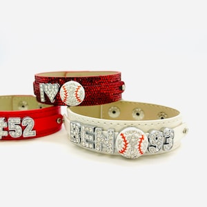 May include: Three leather bracelets with rhinestones and baseball charms. The bracelets are red, white, and black. The red bracelet has the text "#52" on it. The white bracelet has the text "BEN 93" on it. The black bracelet has a heart with the text "I" and a baseball charm.