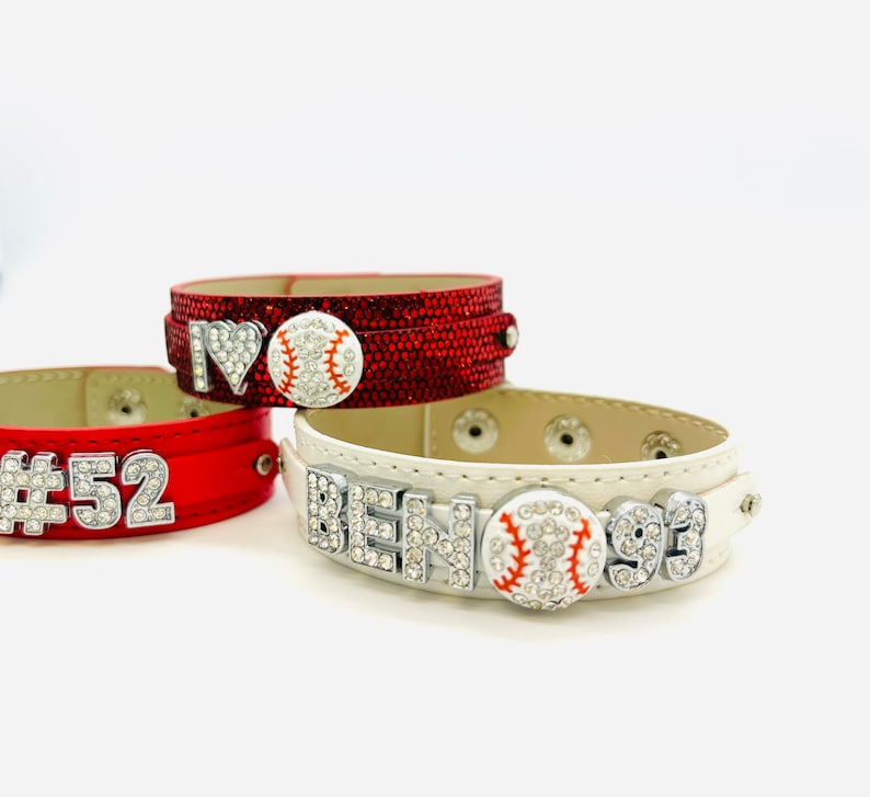 May include: Three leather bracelets with rhinestones and baseball charms. The bracelets are red, white, and black. The red bracelet has the text "#52" on it. The white bracelet has the text "BEN 93" on it. The black bracelet has a heart with the text "I" and a baseball charm.