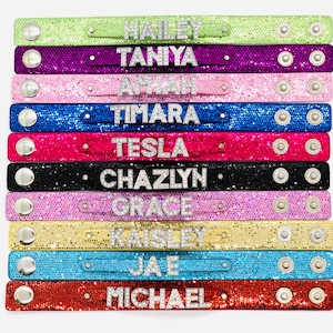 May include: Ten colourful bracelets with different names spelled out in rhinestones. The bracelets are made of a sparkly material and have silver snaps.