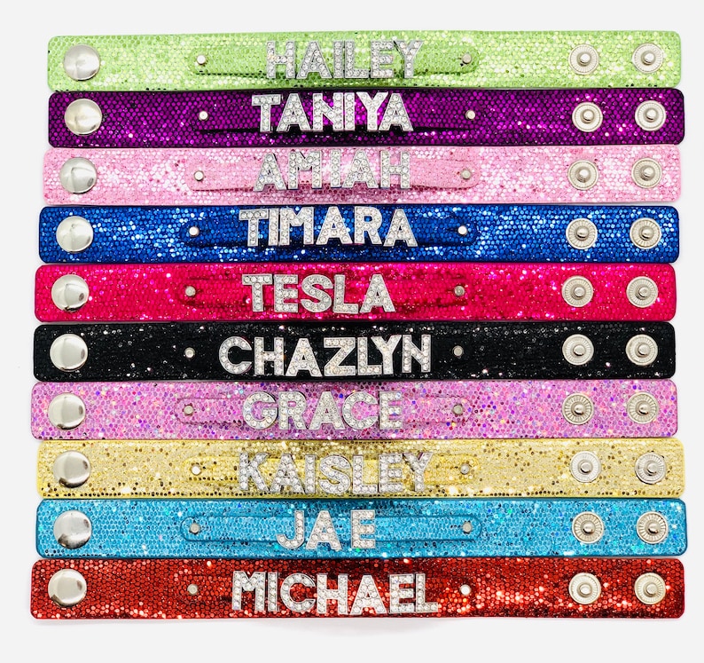 May include: Ten colourful bracelets with different names spelled out in rhinestones. The bracelets are made of a sparkly material and have silver snaps.
