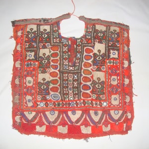 May include: A colourful, hand-stitched textile with a square shape and a neck opening. The piece features a red background with intricate embroidery in white, orange, blue, and green. Small mirrors are incorporated into the design, adding a reflective element.