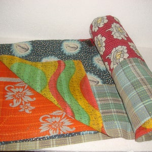 May include: A colorful kantha quilt made from recycled saris. The quilt is made up of several different fabrics, including a red floral print, a green and yellow striped fabric, and a blue and white floral print. The quilt is rolled up and tied with a blue ribbon.