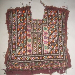 May include: A brown fabric garment with intricate embroidery and colorful beads. The garment is decorated with small mirrors and has a scalloped neckline.