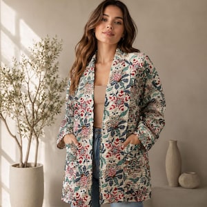 Puede incluir: Una chaqueta acolchada de manga larga y frente abierto con una base color crema y un patrón floral repetitivo en tonos rojos, azules y verdes. La chaqueta incluye dos bolsillos delanteros y se usa sobre una blusa beige y jeans azules. El texto dice EcoFriendlyStuff.