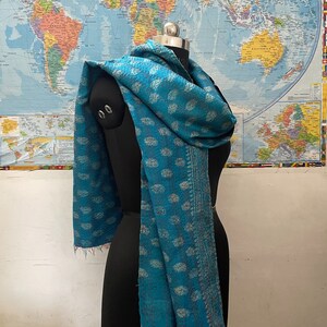 K&ouml;nnte beinhalten: Ein blaugr&uuml;ner Schal mit einem sich wiederholenden Paisley-Muster in einem helleren Farbton. Der Stoff ist durchgehend mit sichtbaren Steppstichen im Kantha-Stil versehen und hat eine dekorative Kante sowie ausgefranste Enden am Saum.