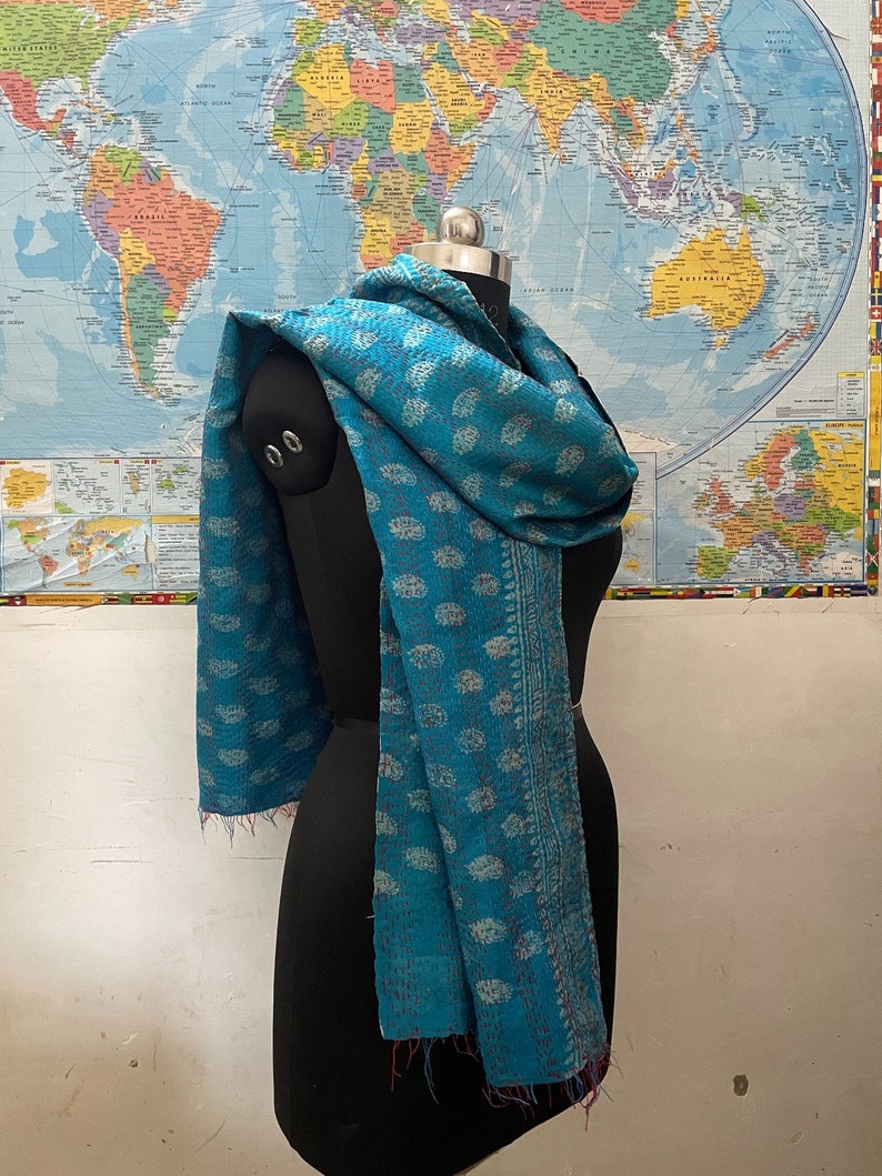 K&ouml;nnte beinhalten: Ein blaugr&uuml;ner Schal mit einem sich wiederholenden Paisley-Muster in einem helleren Farbton. Der Stoff ist durchgehend mit sichtbaren Steppstichen im Kantha-Stil versehen und hat eine dekorative Kante sowie ausgefranste Enden am Saum.