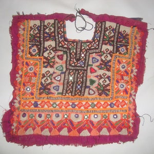May include: A vintage, hand-embroidered textile with a vibrant design. The garment features a deep magenta border and intricate patterns in orange, brown, white, and blue. Small mirrors are incorporated into the design, adding a reflective element. The textile has a fringed edge.