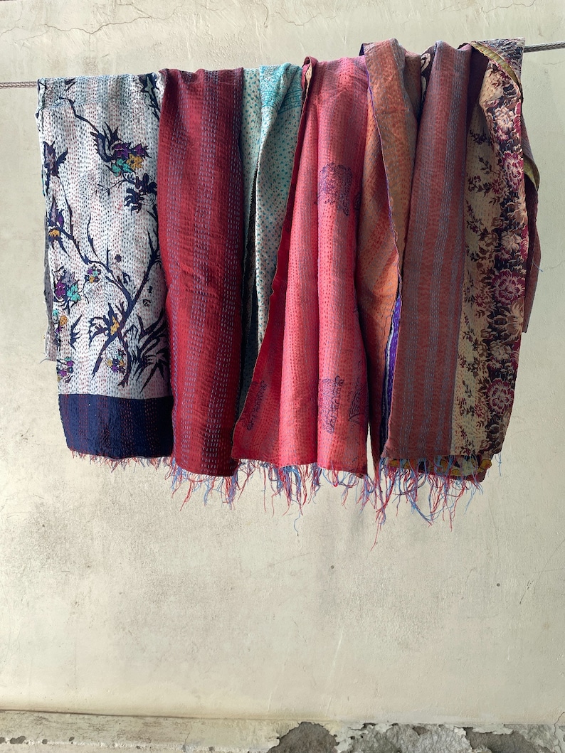 K&ouml;nnte beinhalten: Eine Sammlung rechteckiger Stoff&uuml;berw&uuml;rfe mit komplexen Kantha-N&auml;hten und verschiedenen Mustern, darunter florale Motive, geometrische Designs und kr&auml;ftige Rott&ouml;ne, die mit ausgefransten Kanten versehen sind und an einer horizontalen Leine h&auml;ngen.