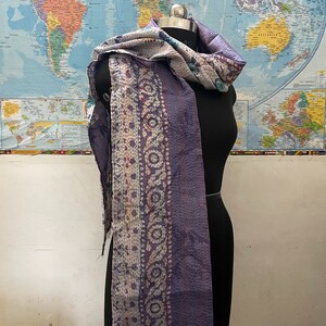 K&ouml;nnte beinhalten: Ein rechteckiger Schal aus einem Patchwork aus violetten, wei&szlig;en und blauen Stoffen mit aufwendigen Kantha-Stickereien. Der Schal zeigt eine Vielzahl von Blumen- und geometrischen Mustern mit sichtbaren ausgefransten Kanten und einer gesteppten Textur.