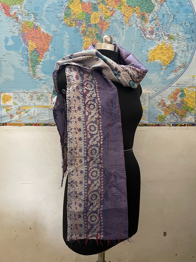 K&ouml;nnte beinhalten: Ein rechteckiger Schal aus einem Patchwork aus violetten, wei&szlig;en und blauen Stoffen mit aufwendigen Kantha-Stickereien. Der Schal zeigt eine Vielzahl von Blumen- und geometrischen Mustern mit sichtbaren ausgefransten Kanten und einer gesteppten Textur.