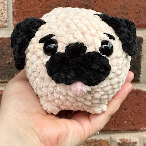 May include: A handmade, plush pug dog with a cream-colored body, black ears and snout, and large black eyes. The pug has a small pink tongue sticking out. The toy is held in a hand against a brick background.