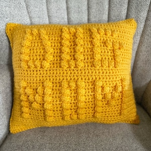 May include: A yellow crocheted pillow with a textured, bumpy design. The pillow is sitting on a gray armchair.