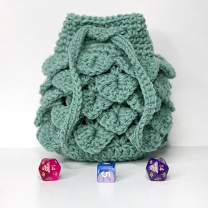 May include: A green crocheted dice bag with a drawstring closure. Three dice are in front of the bag, one pink, one blue, and one purple.