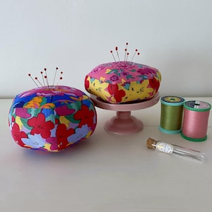 Könnte beinhalten: Zwei bunte florale Stoff-Nähkissen mit Nadeln, eines auf einem rosa Sockel, und zwei Garnrollen, eine grüne und eine rosa, sowie ein Glasröhrchen mit Nadeln.