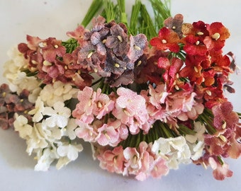 以下が含まれることがあります： ピンク、赤、白の様々な色合いの小さな造花が束ねられています。花は集まっており、繊細でベルベットのような質感があります。