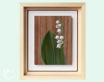 May include: A framed painting of a single lily of the valley flower with white blossoms and a green leaf on a wood background.