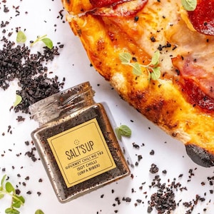 May include: A small glass jar filled with dark red-brown spice labelled "SALT'S UP GOURMET CHILI NO 152 ISOT CHILLI (URFA BIBER)". The jar is sitting on a white surface with a pizza slice in the background.